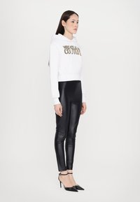 Felpa bianca corta con "Versace Jeans Couture" in oro, abbinata a leggings neri lucidi e tacchi neri a punta.