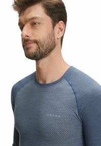 Camicia blu a maniche lunghe con un motivo geometrico testurizzato sul davanti, maniche blu a tinta unita in contrasto e scollatura arrotondata.