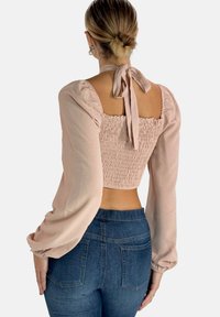 Top corto de color rosa pálido con cuerpo fruncido, mangas largas y voluminosas, y detalle de lazo en la espalda, combinado con jeans oscuros de tiro alto.