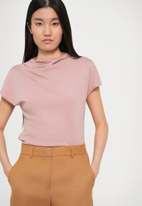 T-shirt ajusté à manches courtes en tissu rose clair avec un col drapé, associé à un pantalon marron sur mesure, doté de poches latérales.