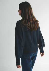 Námorn�ícka oversize pletený sveter s textúrovaným chrbtom, zakončenými manžetami a voľným strihom, v kombinácii s modrými džínsami na neutrálom pozadí.