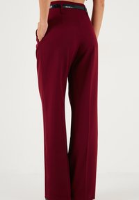 Pantaloni largi burgundy cu o textură netedă, având buzunare laterale și un detaliu lucios în jurul taliei de culoare neagră.