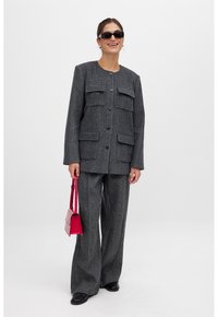 Grijze wollen blazer met vier voorzakken, knoopsluiting en lange mouwen. Gecombineerd met bijpassende wijde broek en een kleine rode handtas.