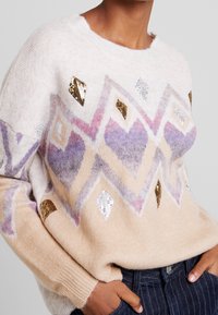 Beige sweater featuring a geometric pattern with purple, gold, and silver accents. Made of soft, textured fabric with sequined details.