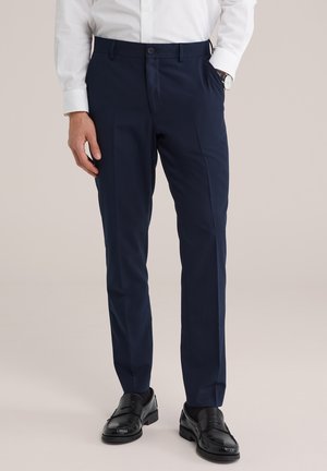 Homme portant un pantalon sur mesure bleu marine, une chemise blanche habillée, des mocassins en cuir noirs et une montre-bracelet, debout avec une main dans la poche.