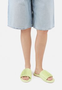 Chanclas deslizantes plisadas de color verde claro con suelas planas, que cuentan con un material suave y un diseño de punta abierta, combinadas con pantalones cortos de mezclilla.