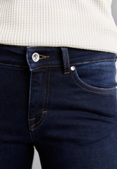 Donkerblauwe denim jeans met oranje stiksels, een metalen knoopsluiting en zijzakken. Geribbelde witte sweater zichtbaar aan de bovenkant.