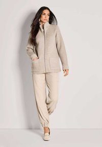 Frau mit langen dunklen Haaren, die eine beigetönige, gerippte Strickjacke mit Reißverschluss, passende lockere Hose und helle Slipper trägt und vor einem schlichten Hintergrund steht.