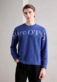Sudadera azul con gran texto blanco "Marc O’Polo", con un clásico cuello redondo y puños acanalados, combinada con pantalones negros.