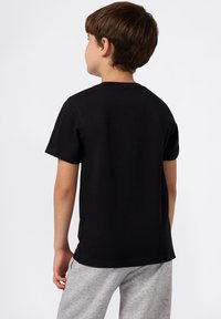 Ragazzo con i capelli castani che indossa una semplice maglietta nera a maniche corte e pantaloni grigi, in piedi di spalle contro uno sfondo bianco.