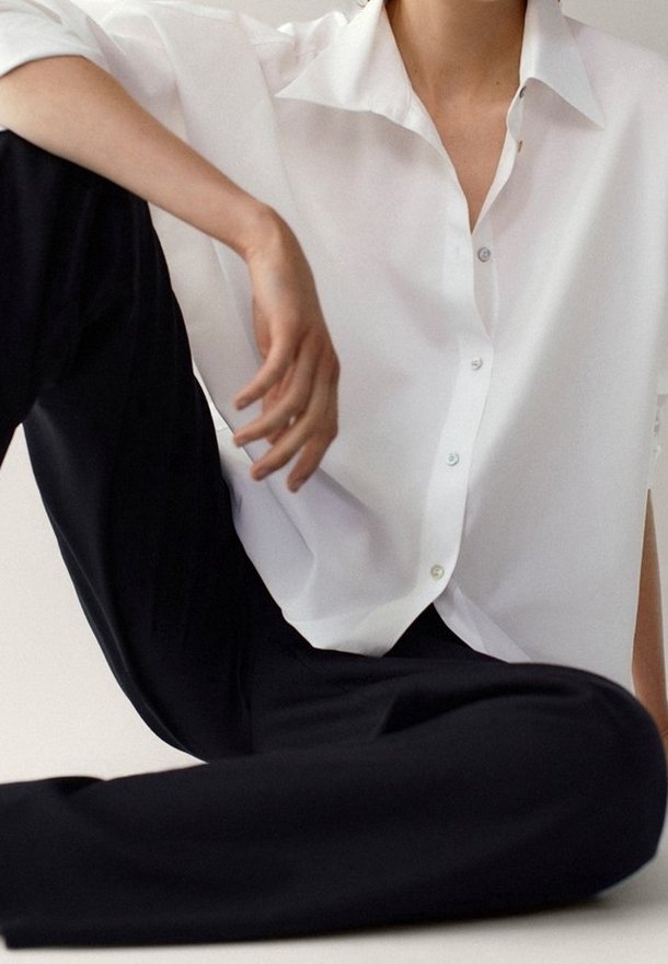 Personne assise en tailleur portant une chemise blanche ample à boutons et un pantalon noir, un bras reposant sur un genou plié.