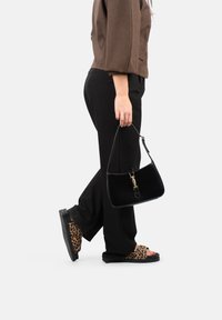 Borsa in velluto nero con una chiusura dorata, portata da una persona che indossa pantaloni neri e sandali con stampa leopardata.