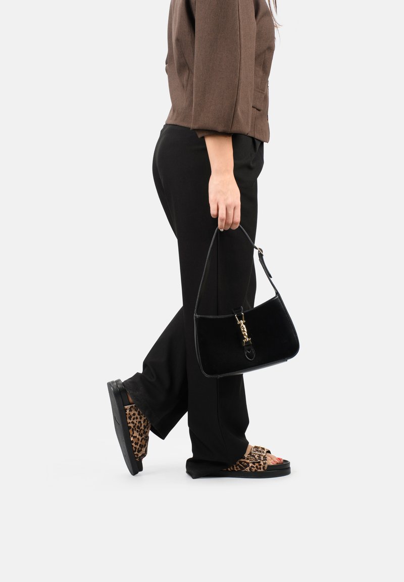 Borsa in velluto nero con una chiusura dorata, portata da una persona che indossa pantaloni neri e sandali con stampa leopardata.