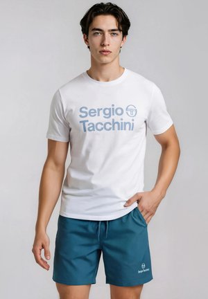 Ung mand står iført en hvid Sergio Tacchini t-shirt og blå shorts med en hånd i lommen mod en ensfarvet baggrund.
