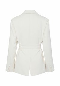 Hvid blazer med en bælte i taljen, udskåret reverskrave og lange ærmer. Har en slids på bagsiden og knapdetaljer på ærmerne.