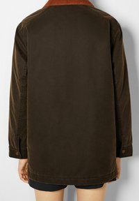 Brun, voksbehandlet bomuldsjakke med ribbet krave. Har lange ærmer, trykknapper og lige snit. Bagfra ses minimal detaljering.