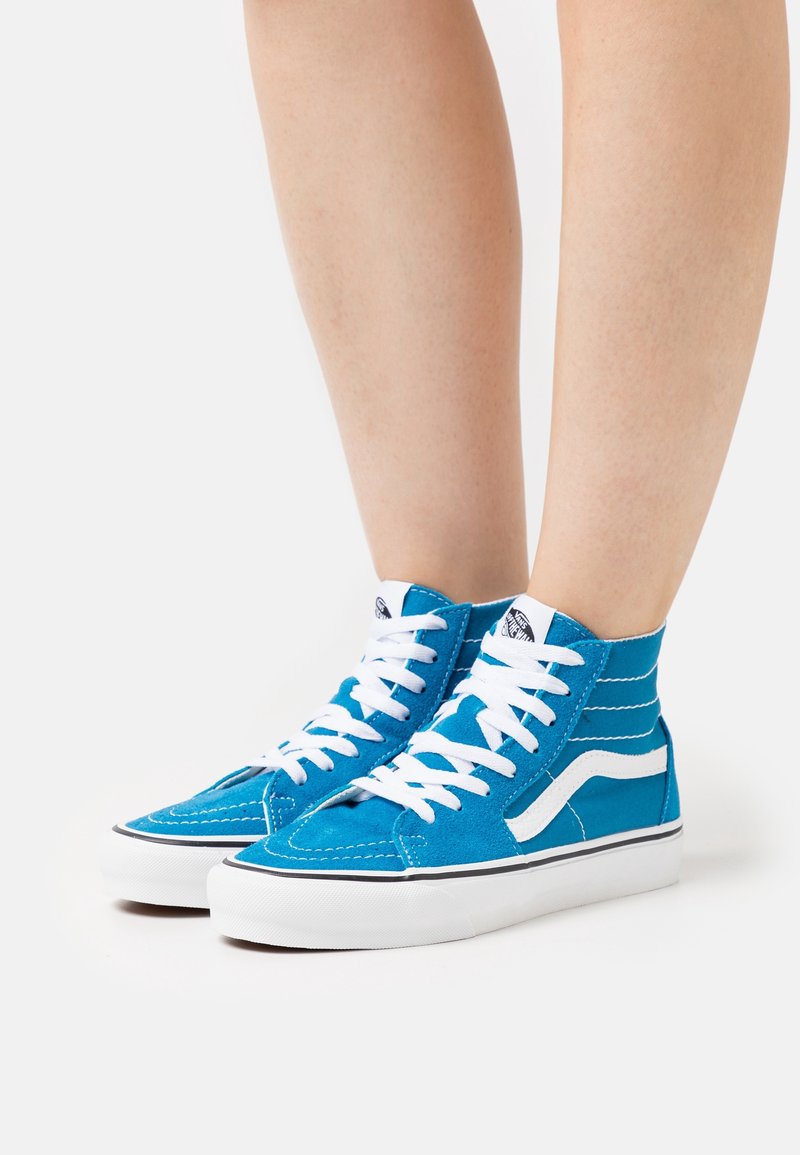 Jambes portant des baskets montantes bleues avec des lacets blancs et une semelle blanche, debout sur un fond blanc.