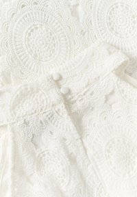 Tissu en dentelle blanche avec des motifs circulaires au crochet et deux boutons recouverts de tissu sur un col à bord festonné.