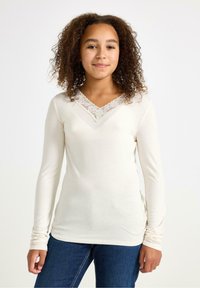 Giovane con capelli ricci che indossa una maglietta a maniche lunghe color crema con bordino in pizzo e jeans blu, in piedi contro uno sfondo bianco uniforme.