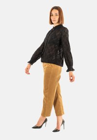 Blusa di pizzo nero con maniche lunghe, chiusura anteriore a bottoni e colletto alto, abbinata a pantaloni di velluto a coste color cammello e scarpe nere con tacco alto.