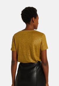 T-shirt à manches courtes en tissu doré métallisé, avec une coupe décontractée, un col en scoop et une surface douce et texturée. Associé à une jupe noire.