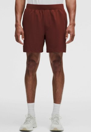 Partie inférieure du corps d’un homme portant un short de sport rouge foncé, des chaussettes blanches montantes et des chaussures de course blanches, debout droit sur un fond uni.