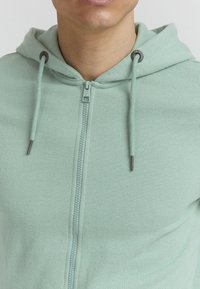Lichtgroene zip-up hoodie met een gestructureerde stof, voorzien van een trekkoordkap en donkere metalen accenten op de rits en trekkoorden.
