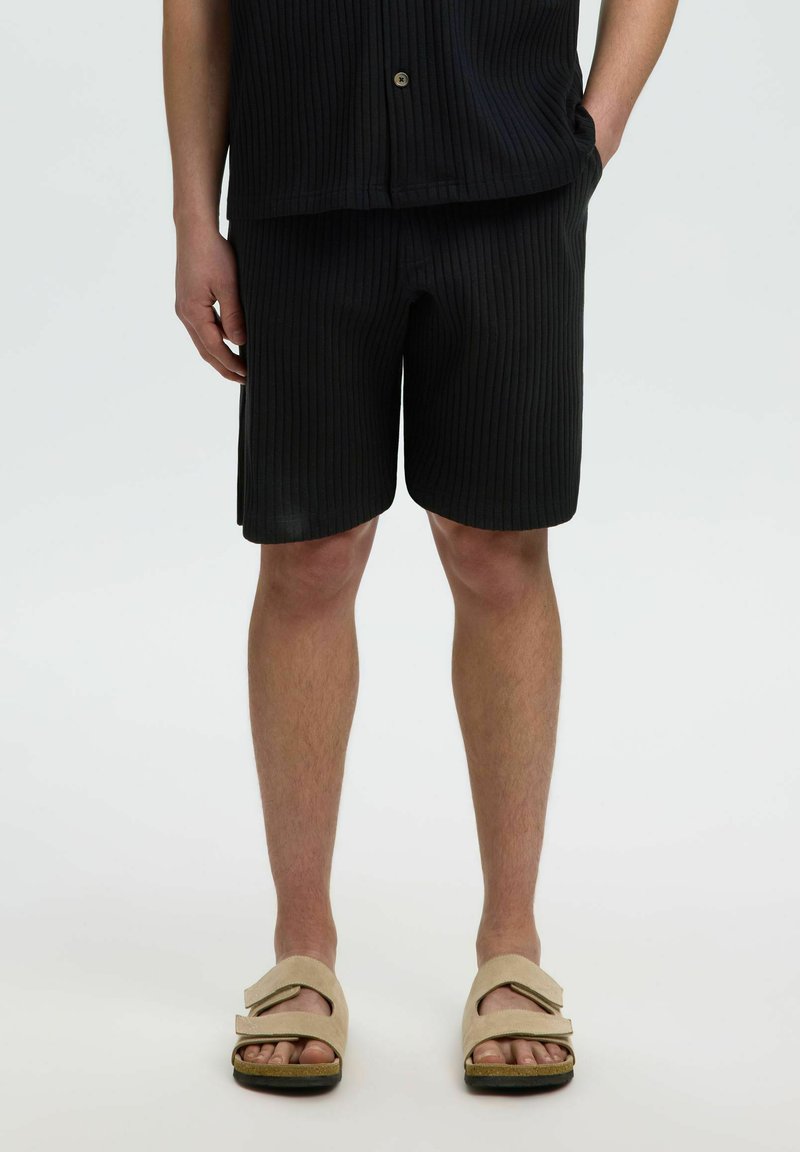 Homme portant un short noir côtelé et une chemise assortie, des sandales beige à brides, debout sur un fond clair, les mains dans les poches.