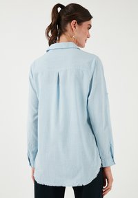 Camicia a maniche lunghe azzurro chiaro con tessuto testurizzato, dettaglio delle pieghe sul retro e orlo curvo. Presenta polsini con bottone e colletto classico.