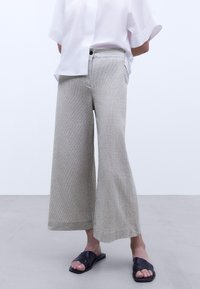 Lichtgrijze wijde broek van textuurstof, met een hoge taille, voorknoop en zijzakken, gecombineerd met zwarte sandalen.