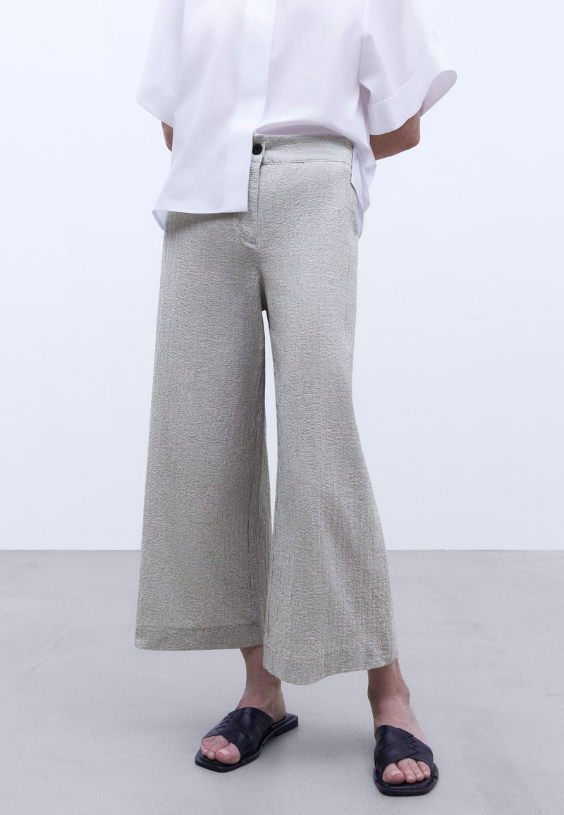 Lichtgrijze wijde broek van textuurstof, met een hoge taille, voorknoop en zijzakken, gecombineerd met zwarte sandalen.