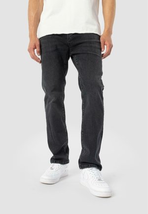 Person wearing black jeans and white sneakers standing against a light background, with hands relaxed at sides.