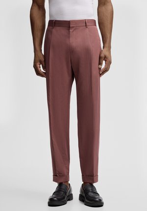 Man draagt op maat gemaakte stoffige rozetinten broek met omslagen en zwarte leren loafers, staand tegen een effen achtergrond.