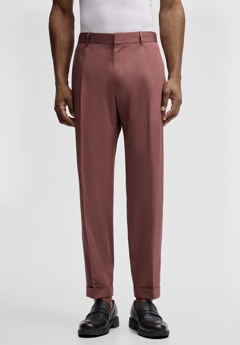 Homme portant un pantalon sur mesure rose poussiéreux avec revers et des mocassins en cuir noir, debout devant un fond uni.