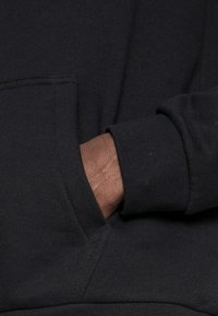 Svart huvtröja med en framficka som har känguru-design och ribbade ärmslut. Tyget verkar mjukt med en slät textur. En hand är placerad inne i fickan.