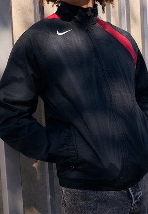 Person trägt eine schwarze Nike Zip-Jacke mit einem roten diagonalen Streifen über der Schulter, steht an einer grauen Wand mit den Händen in den Taschen.