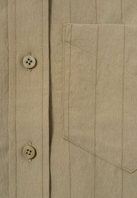 Tan skjorta med knappar, vertikal söm, bröstficka och fyra knappar med plastknappar. Slät textur.