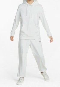 Persona con sudadera blanca y pantalones de chándal blancos sueltos, vistiendo zapatillas blancas, de pie frente a un fondo blanco liso.