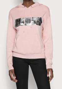 Persona che indossa una felpa leggera rosa con un brillante logo "GUESS" argento sul petto e pantaloni neri.