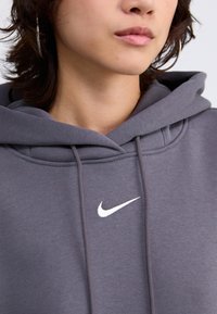 Szürke kapucnis pulóver puha anyagból, elülső zsebbel, húzózsinórral ellátott kapucnival és fehér Nike logóval a mellkasán.