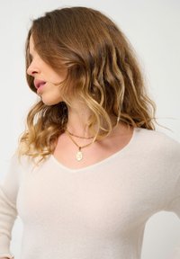 Maglione beige chiaro con scollatura a V, caratterizzato da una texture morbida. Collane in oro a strati con un pendente dal design profilato. Capelli ondulati visibili.