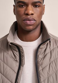 Beige gesteppte Jacke mit hohem Kragen und schwarzem Reißverschluss, kombiniert mit einem hellen, gerippten Pullover. Dezentes Branding am Kragen.