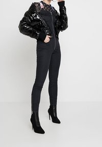 Černá zimní bunda s lesklým povrchem, spárovaná s černými kombinézami a vysokými botami na podpatku. Tkanina celkových kalhot je hladká; zipy na kalhotách jsou viditelné.