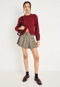 Πλεκτό cardigan σε τούβλινο κόκκινο με κουμπιά, συνδυασμένο με πλισέ φούστα καρό. Καφέ μοκέτες και μαύρες κάλτσες συμπληρώνουν την εμφάνιση.