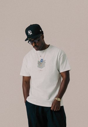 Weißes T-Shirt mit kurzen Ärmeln, das ein Grafikdesign auf der Brust zeigt, kombiniert mit einer schwarzen Kappe und Hose. Goldschmuck ergänzt das Outfit.