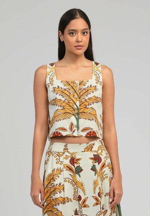 Femme portant un crop top sans manches boutonné et une jupe assortie avec un motif de feuilles jaunes, rouges et vertes sur fond blanc.