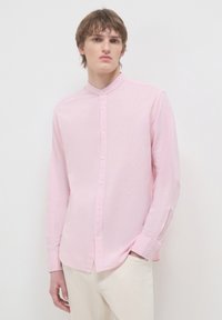 Junger Mann in einem hellrosa, geknöpften Hemd mit Mandarin-Kragen und cremefarbenen Hosen, der vor einem schlichten weißen Hintergrund steht.