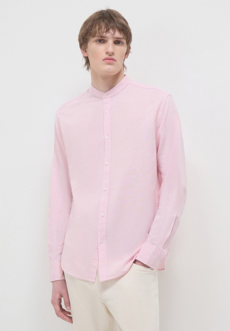 Junger Mann in einem hellrosa, geknöpften Hemd mit Mandarin-Kragen und cremefarbenen Hosen, der vor einem schlichten weißen Hintergrund steht.