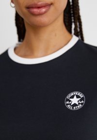 Μαύρο κοντομάνικο μπλουζάκι με λευκό ραμμένο γιακά, που έχει έναν στρογγυλό κεντητό λογότυπο 'Converse All Star' στο στήθος.