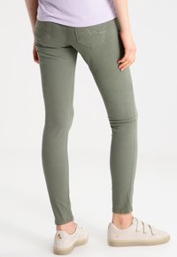 Jeans skinny elásticos de color verde oliva con una textura suave, con bolsillos traseros y costuras decorativas, combinados con zapatillas de color claro.